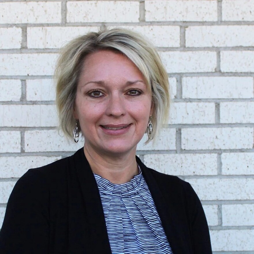 Headshot of a pretty blonde woman. She is wearing a black and white thinly striped shirt and a black blazer.
