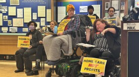 Janine Hunt-Jackson (middle, yellow hat) watches as Caring Majority activist Renee Christian speaks into a microphone at a rally at Buffalo and Erie County's Central Library. Both are using power wheelchairs. They are surrounded by other activists holding signs.