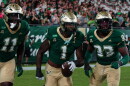 3 football players wearing green jerseys and gold helmets and pants running together, with player in middle carrying football in left hand
