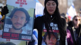 Woman wearing black beanie hat holds sign of young man with the words "Bring Him Home Now." She wears a T-shirt with same image.