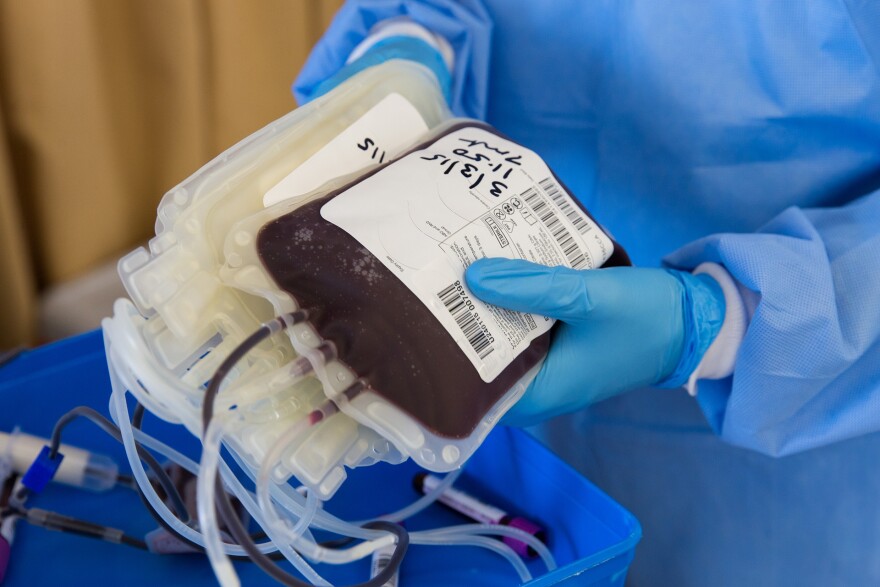 A person in gloves and medical gown holding an IV bag of blood.