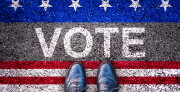 Feet on and asphalt road with the word vote. Red and white stripes are below the word. White stars on a blue stripe are above.
