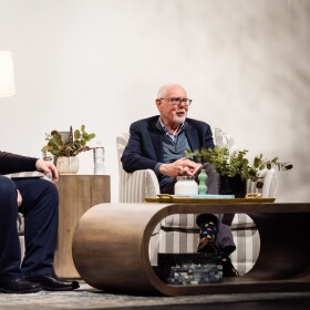 Tom Williams and Corey Flintoff, two older white men, sit on a stage during an audience Q&A.