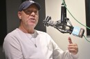 A man wearing a baseball cap and white shirt sits inside a radio studio. He is gesturing with his left hand and talking animatedly at a microphone.