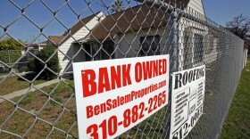 A vacant home surrounded by a chain link fence, carrying a "bank owned" sign.