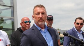 A man in a nice jacket looks serious as he stands behind a microphone. Other men look on. 