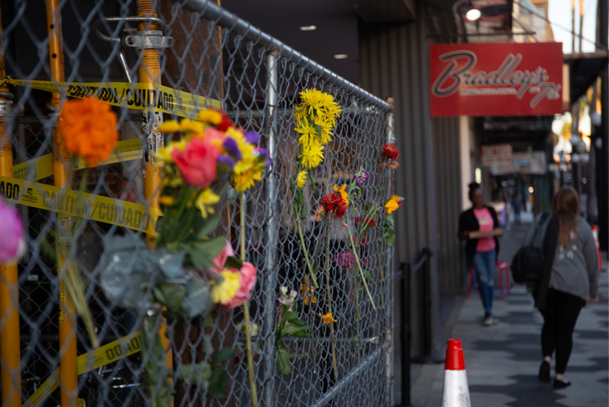 Una valla metálica con cinta de precaución y flores entretejidas. Al fondo, el letrero rojo de Bradleys.