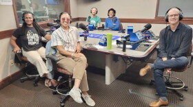 Five smiling people wearing headphones sit around a table in a radio talk studio: A woman front left has long black braids and is wearing white pants, black shoes, and a black graphic t-shirt with white text that reads "STREETS ARE FOR PEOPLE;" a woman front center has her dark hair tied back and is wearing sunglasses, a blue and white collared button-down shirt, tan pants, and white sneakers; a man back left is bald and is wearing a green graphic t-shirt; a woman back center has short black hair and is wearing a blue top; a man at right has very short dark hair and is wearing a black button-down shirt, jeans, and brown sneakers