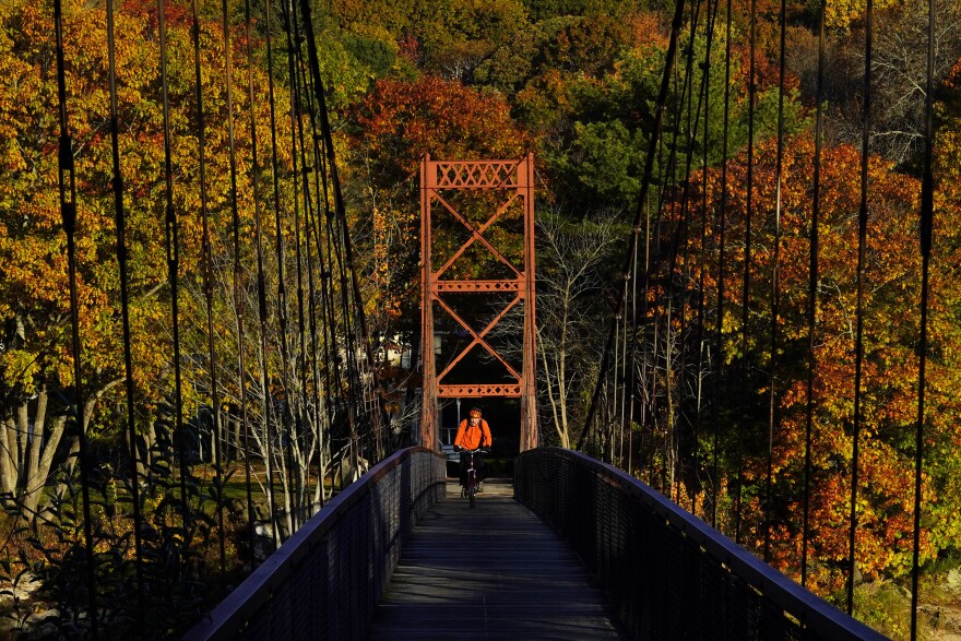 Fall foliage provides a colorful backdrop as a bicyclist rides across the Swinging Bridge spanning the Androscoggin River, Oct. 29, 2021, in Brunswick, Maine. 