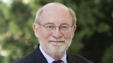 A bald man in a suit and dark purple tie looks into the camera. He is wearing glasses, a pin is on his right lapel. 
