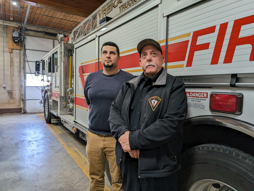 Two men stand in front of a large, white and chrome fire truck with red and golden accents. The men on the left wears a black jacket with a logo that says, "Yreka Fire Dept." They are looking into the distance.