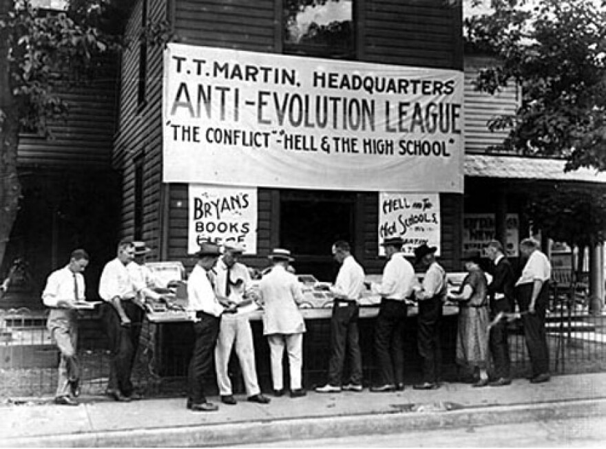 Black-and-white photo of a group of men in suits standing around a table underneath a sign reading, "T.T. Martin. Headquarters. Anti-Evolution League. 'The Conflict' – 'Hell and the High School'"