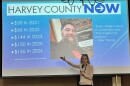 A woman stands in front of a PowerPoint screen with her hand extended, speaking into a microphone. An image of a bearded man and some info about “Harvey County Now” is displayed behind her.
