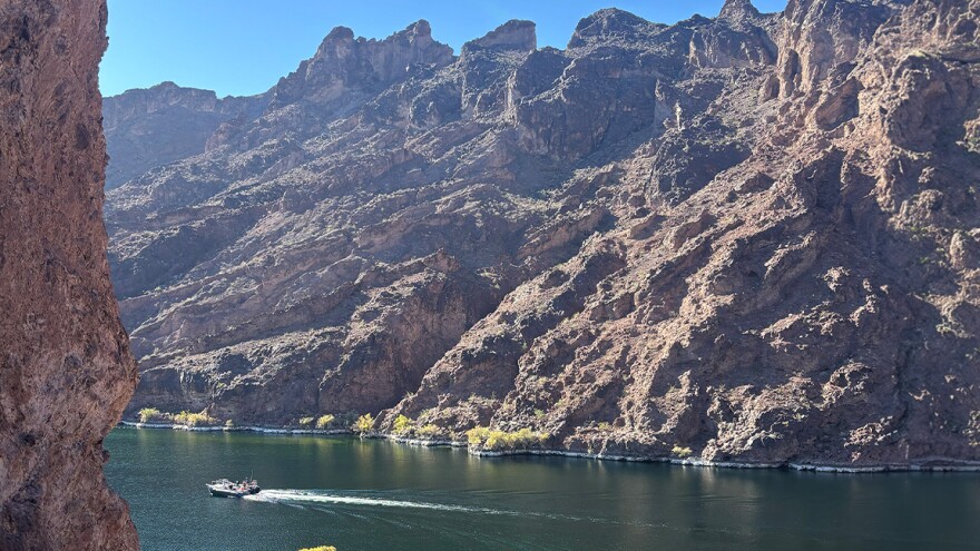 A small motorboat cuts through the calm, dark green waters of the Colorado River, flanked by towering dark volcanic canyon walls under a clear blue sky, with sparse yellow-leafed trees along the shoreline.