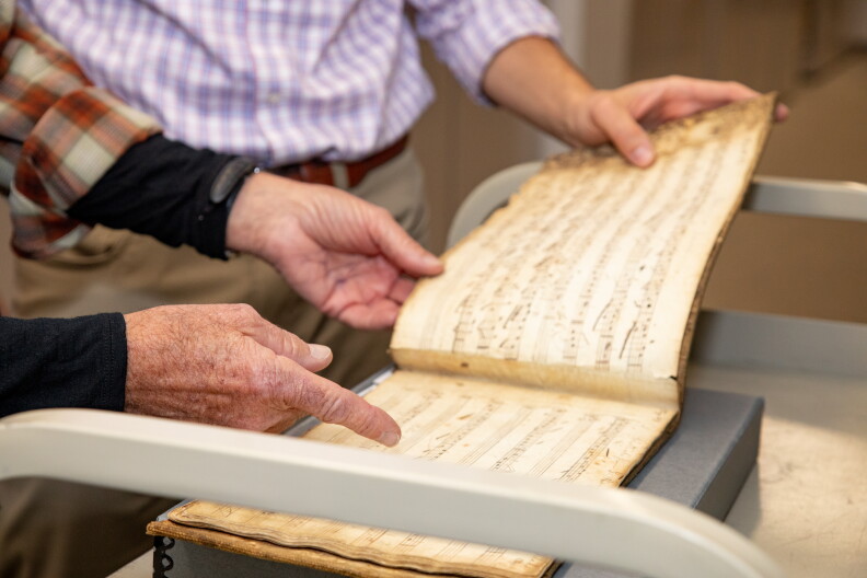 A set of hands points at an old book with yellow pages full of musical notation.
