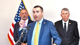 Man in blue suit and yellow tie stands at podium speaking 