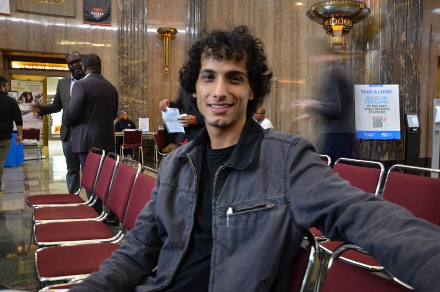 A man in a grey jacket sits in a red chair, facing the camera