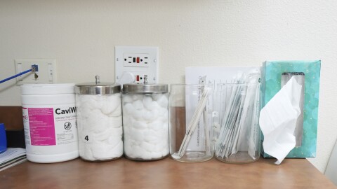 Cotton balls in jars, a box of tissues set with the bottom against the all, and other medical supplies are seen on a brown countertop