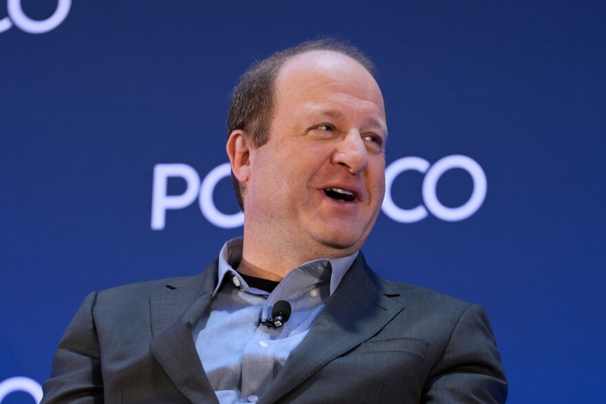 Gov. Jared Polis wears a suit and speaks in front of a blue sign that has the Politico logo on it.