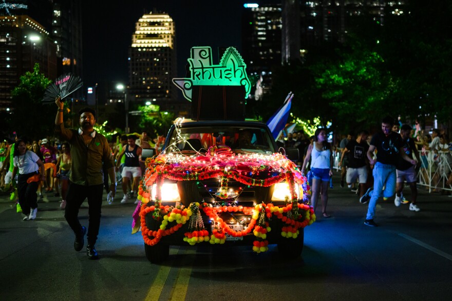 A truck lit up with lights and colorful balls with the KhushATX logo in the back makes its way down Congress Avenue during the Austin Pride Parade. People walk along side it waving with crowds lining the street. Lit-up skyscrapers are seen in the background.