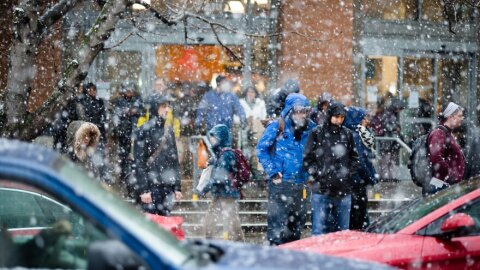 Snow falls as cars and people move across a street.