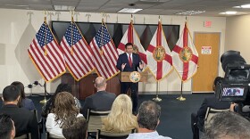 A man in a suit stands at a podium flanked by American flags and the flag of the State of Florida.