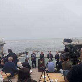 Six nicely-dressed dignitaries stand along a glass fence with Monterey Bay churning behind them. In the foreground, on a slope with bench seats, a handful of people sit with notebooks and cameras. 