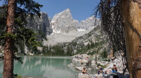 A rocky granite peak rises over an aquamarine lake. People sit and stand on rocks near the shore.