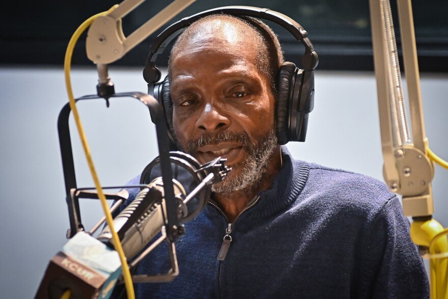  A man wearing a blue shirt talks at a microphone inside a radio studio.