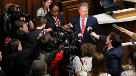 Todd Huston stands in front of a crowd of reporters holding microphones and recorders up at him. Huston is a White man with dark hair. He is wearing a suit and tie.