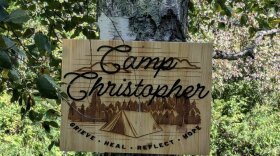A wooden sign reading "Camp Christopher: Grieve, Heal, Reflect, Hope" is on a birch tree with vegetation in the background. 