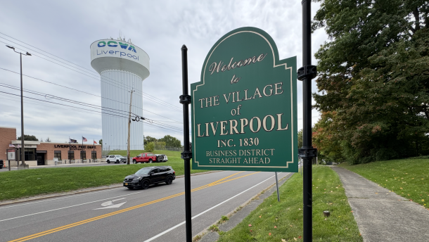 A green sign with gold lettering stands across a two lane road from a Liverpool firestation and water tower. The sign reads, "Welcome to The Village of Liverpool Inc. 1830 Business District Straight Ahead."
