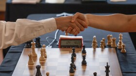 Two hands shake over a chess board with a red score keeping box at the edge. 