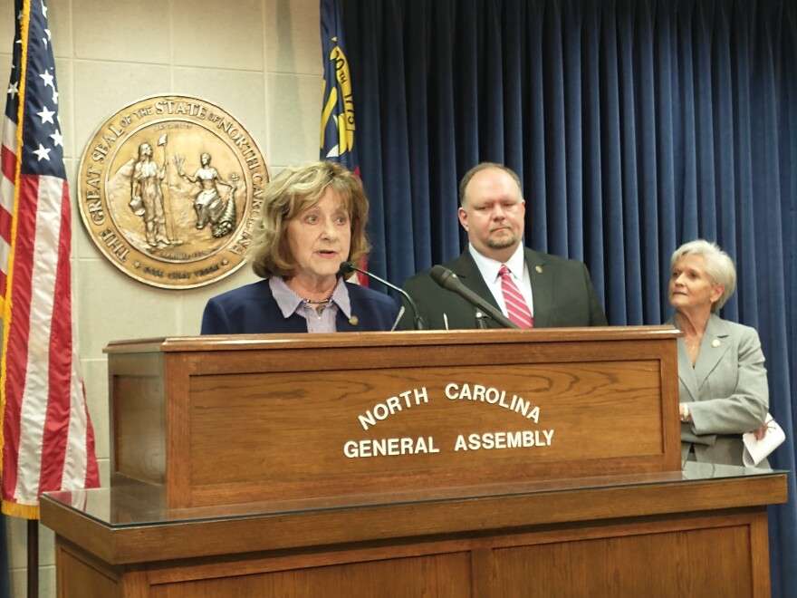 Three people stand by a wood podium. A large metallic seal of North Carolina and American flags are in the background.
