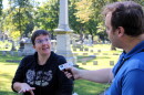 A man with a microphone interviews a woman in a cemetery. 