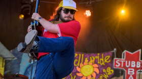 A long-haired bearded man in a trucker hat, cowboy boots, jeans and sunglasses on stage, kicking his leg up and strumming his electric guitar off to his side