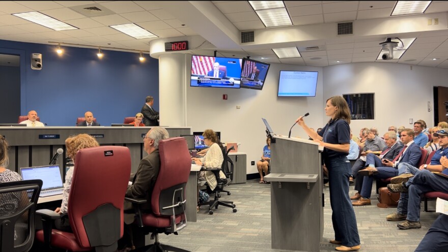 Woman in jeans and blue shirt stands at podium and addresses official sitting behind a dais