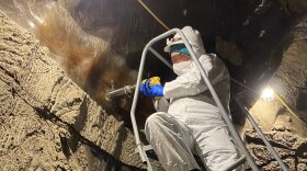 A person in a full-body, white suit with their face covered and protective glasses uses a drill-like piece of equipment to drill into a frozen dirt wall.