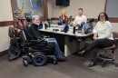 Five people sit at a table in a radio talk studio: a woman at left in foreground has short red hair and is wearing glasses, a red button-down shirt, black t-shirt and black pants; a woman at right in foreground is in a power wheelchair and has short purple hair and is wearing glasses, a black turtleneck, jeans and black shoes; a woman at left in background has long red hair and is wearing a yellow knit cap, a yellow and black plaid flannel button-down shirt and black t-shirt; a man at right in foreground has light brown hair and a light brown beard and is wearing a pink button-down shirt; a woman at center has short dark hair and is wearing glasses, a white sweater, black jeans and black shoes.