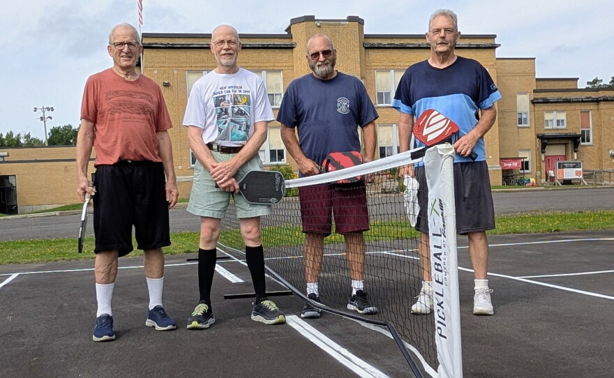 Men stand on both sides of a pickleball net in front of an old school building.