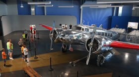 A silver, twin-propeller airplane sits inside a large building. School children and adults stand nearby, looking at it.