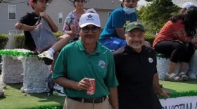 Two men stand in front of a parade float with two young men and a woman sitting on top of it.