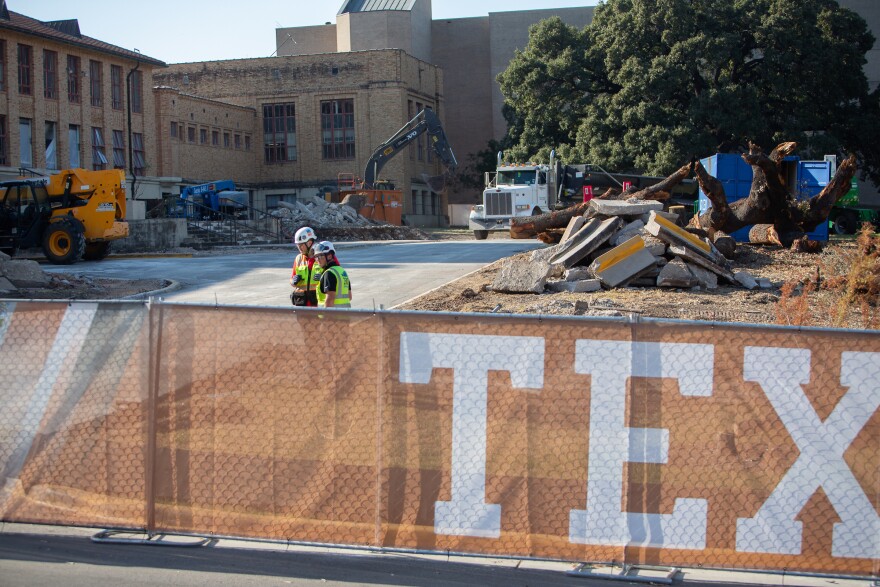 Trabajadores con chalecos amarillos detrás de una valla en una obra con escombros y camiones.