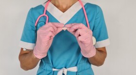A medical worker in blue scrubs and pink latex gloves holds up a heart-shaped pin in the trans flag colors. 