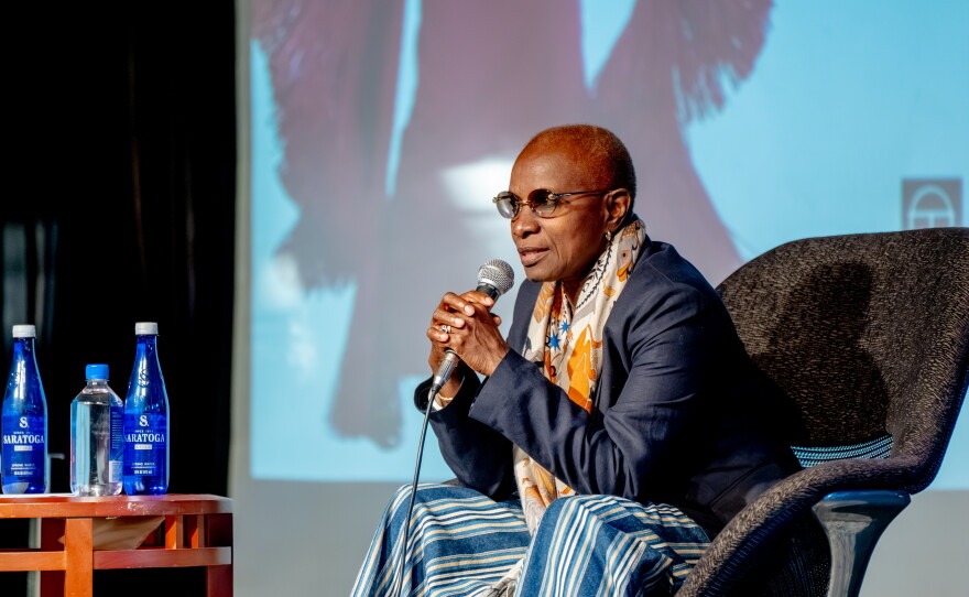 A woman wearing glasses, a dark jacket and a patterned scarf sits onstage holding a microphone during a speaking event.