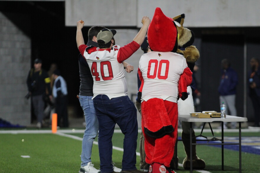 Four people, including a person in a red bird mascot costume and another in a "40" jersey, celebrate together on a football field near a table with pizza boxes. Their backs are facing the camera.
