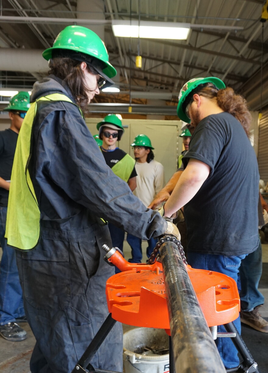 Young people in green hardhats gather around a long pipe in a workshop space.