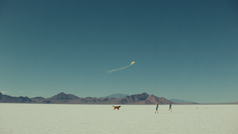 A dog chases after two kids flying a kite, mountains are visible in the background.