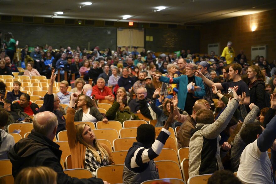 People in a crowd in an auditorium raise their hands to be counted on caucus night 2020. 