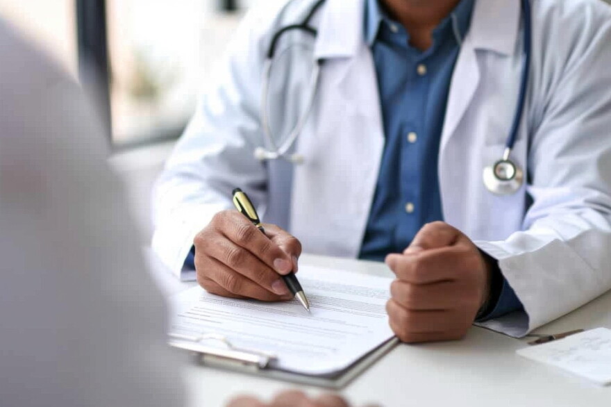 A doctor, with a stethoscope around his neck, writes on a paper on a desk
